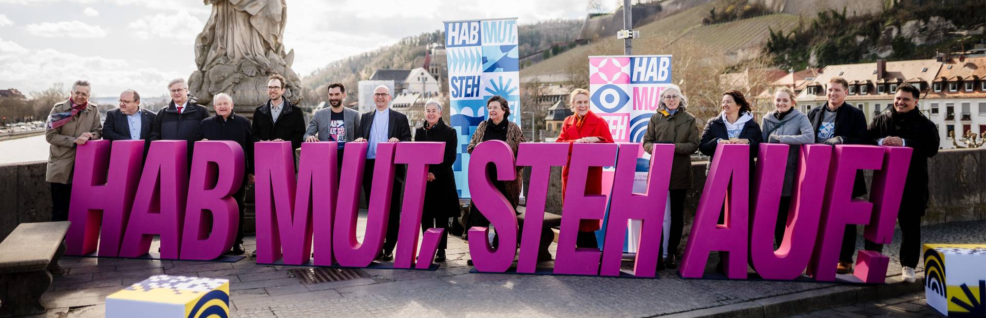 Menschen halten große, pinke Buchstaben mit „HAB MUT. STEH AUF!“ auf Brücke.
